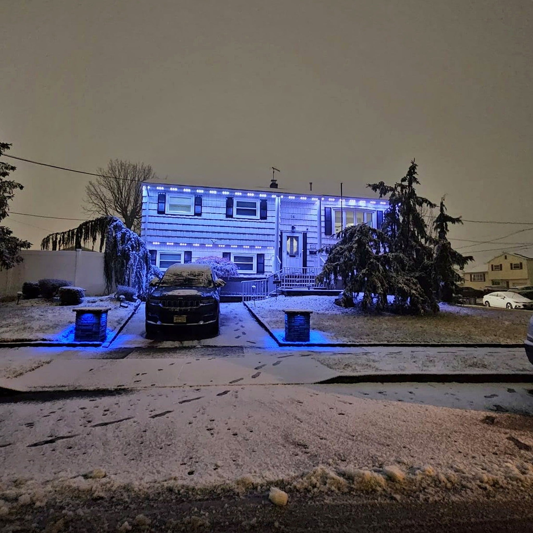 Snow-covered home with blue permanent LED roofline lighting and illuminated driveway pillars