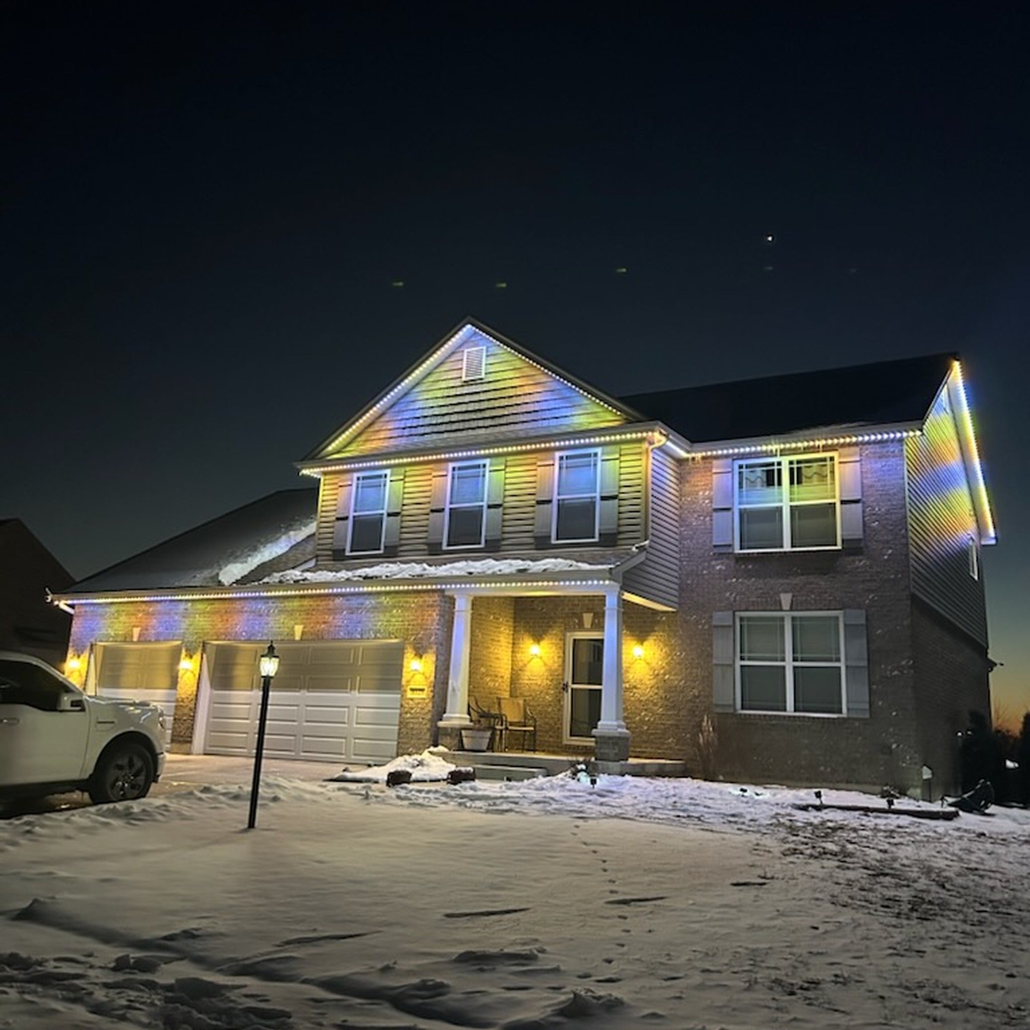 Snow-covered two-story home with multicolor rainbow permanent LED roofline lighting